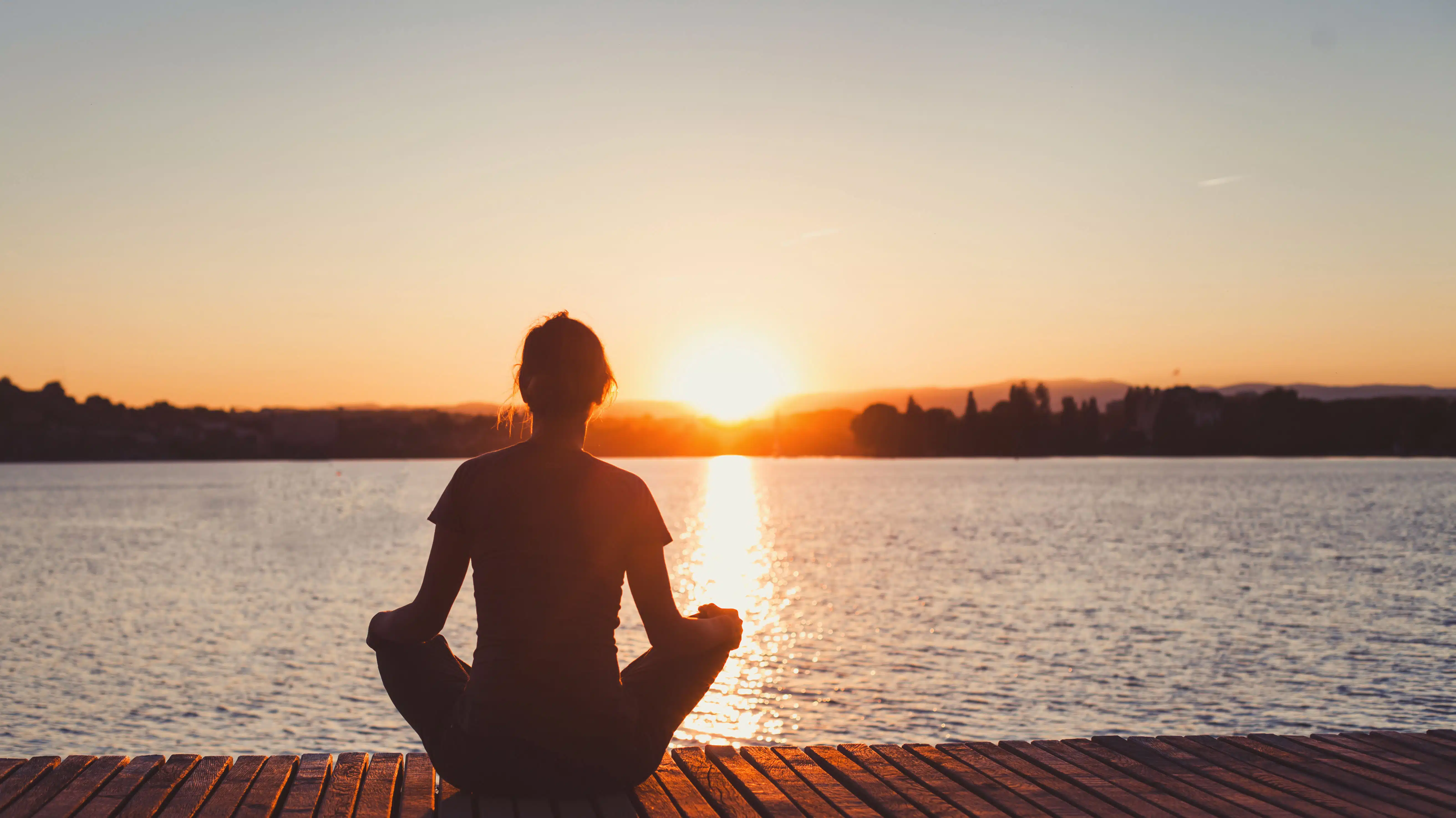 A woman meditates on a dock while the sun sets - 7 Complementary Health Approaches For Addiction
