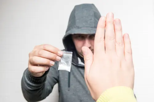 hand seen gesturing "stop" to a hooded man holding up a small bag of cocaine.