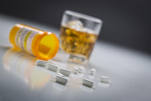 several prescription drugs spilled from a fallen bottle near glass of alcohol.